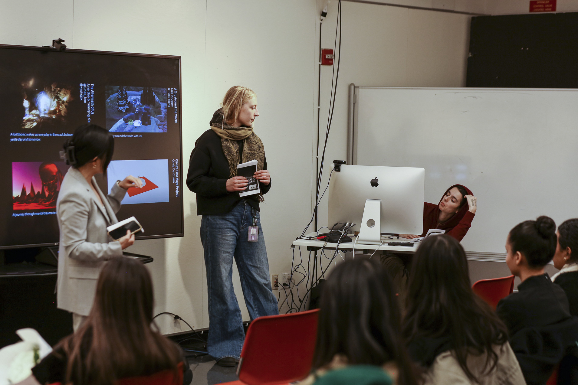 Professor Snow Fu and student Cora Rafe both stand near a television while another student sits in front of a computer and other students are seated nearby